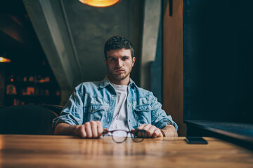 Portrait of confident caucasian male student spending free time and posing at modern university campus, handsome concentrated young hipster guy looking at camera while sitting at wooden table indoors