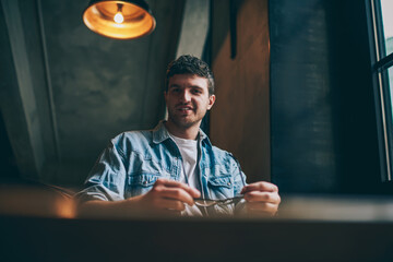 Portrait of handsome successful hipster guy with cute smile on face looking at camera and resting on leisure indoors, positive cheerful male student in stylish jeans shirt enjoying break at campus