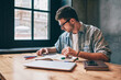 © BullRun - Young concentrated male student making research for course work using modern technology at wifi zone, clever hipster guy reading news about university in break of drawing sketches in notebook