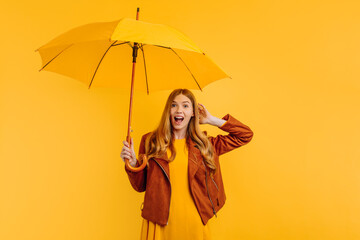 A shocked girl, in a bright yellow dress and an autumn jacket, stands with a yellow umbrella on an isolated yellow background. Autumn concept