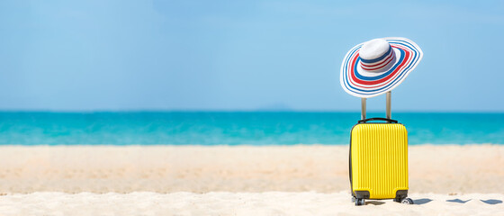 Summer traveling and tourism planning with yellow suitcase luggage with big hat fashion in the sand beach. Travel in the holiday, blue sky and beach background. 