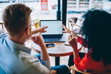 Back view of multicultural business partners pointing on modern laptop computer with blank screen area for your internet content and discussing design of website during collaboration
