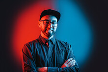 Neon Lights. Portrait Of A Man In A Shirt, Glasses And Cap, With A Beard, Posing With His Arms Crossed. Red And Blue Circle In Black Background