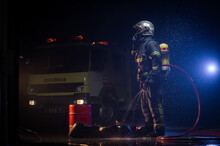 Side View Of Fireman Wearing Protective Uniform And Hardhat Standing With Water Hose During Rainy Night Near Fire Truck And Looking Away