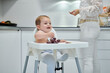 © Gabriel Trujillo/ADDICTIVE STOCK - Cute cheerful toddler sitting in high chair and eating sweet fresh grapes while unrecognizable mother standing nearby in home kitchen