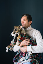 Adult Man In Sweater Looking Away And Embracing Adorable Whippet Dog In Checkered Blanket While Sitting Against Gray Background