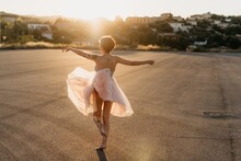 Full Body Of Delighted Unrecognizable Female Artist In Elegant Dance Dress Laughing And Dancing In Back Lit In Empty Square With Blurred Town In Background In Sunny Summer Sunset Day
