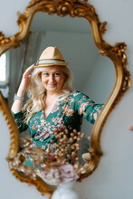 Charming Content Female Wearing Floral Green Dress And Trendy Hat Looking In Cozy Mirror With Golden Frame While Touching Hat