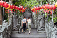 Full Body Cheerful Female Friends In Tight Casual Outfits Strolling Together Along Stone Stairway Decorated With Red Chinese Lanterns In Green Summer Garden