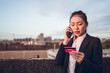 © Juan Alberto Ruiz/ADDICTIVE STOCK - Serious young Asian businesswoman in formal wear holding credit card and having phone conversation with bank customer service while standing on rooftop of city building