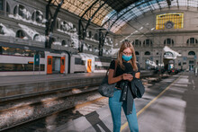 Young Woman Wearing A Face Mask Waiting At The Train Station And Looking At The Smartphone During A Pandemic
