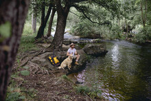 Young Man In A Hat And Yellow Backpack Sitting In The Shade On The Shore Of A Tree-filled Lake Lounging With His Beige Dog After Hiking