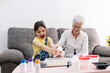 © Sergio Victor Vega/ADDICTIVE STOCK - Smiling granny in eyeglasses helping positive granddaughter with dirty face in casual wear preparing blend using colorants in plastic container while sitting on comfortable couch in living room