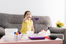 Child In Yellow T Shirt With Spoon Adding Purple Substance Into Rectangular Plastic Container While Preparing Blend And Sitting On Couch In Living Room