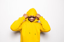 Cheerful Black Man Over White Plain Background. He Is Looking At Camera And Smiling Wearing A Yellow Hoodie. He Is Covering His Face With The Hood.