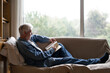 © Westend61 - Senior man reading book while relaxing on sofa by window at home