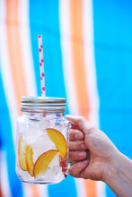 Close-up Of Woman's Hand Holding Drink In Mason Jar Filled With Ice And Slices Of Peach