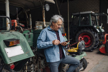 Senior Man Using Tablet On A Farm With Tractor In Barn