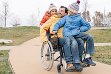 Happy Man With Playful Sons Riding Wheelchair On Footpath At Park