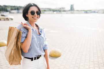 Shopping time. Trendy beautiful woman is happy with shopping. A young caucasian smiling woman in a stylish wear and sunglasses holds paper shopping bags in hand and smiling