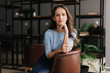 © dikushin - Beautiful young businesswoman wearing stylish light blue dress sitting at the chair in office with modern interior, looking at the camera. Portrait of charming business lady.
