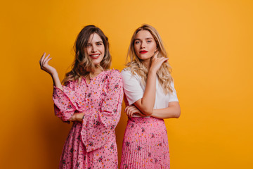 Blithesome white girl in dress relaxing with best friend. Indoor shot of glad caucasian sisters isolated on yellow background.