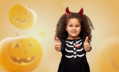 Wall Mural - halloween, holiday and childhood concept - smiling african american girl in black costume dress and red devil's horns over jack-o-lanterns on orange background