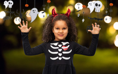 Wall Mural - halloween, holiday and childhood concept - smiling african american girl in black costume dress and red devil's horns over garland and festive lights background