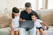 © Mangostar - Two happy kids and their dad using laptop while sitting on couch at home, staring at display. Medium shot, front view. Communication or leisure at home concept