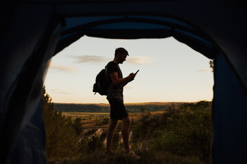 View from tent of man traveller with backpack holding mobile phone. Young man camping on the mountain, standing near tent  and typing a text message.