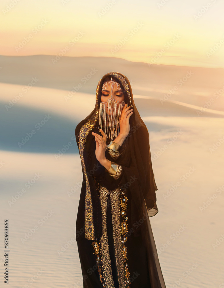 Beautiful mysterious arab woman Queen in black, muslim dress. head with  silk handkerchief, golden chains hides face. A Fantasy Girl oriental  princess walks in desert. art background yellow sand sunset Stock Photo |