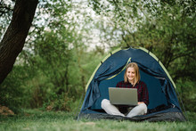 Woman Working On Laptop In Tent In Nature. Young Freelancer Sitting In Camp. Relaxing In Camping Site In Forest, Meadow. Remote Work, Outdoor Activity In Summer. Happy Girl Relaxing, Work On Vacation.