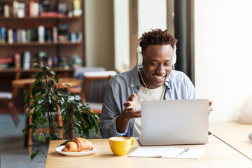 Happy black guy in headphones communicating online from laptop computer at coffee shop, copy space