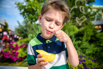 Wall Mural - Tasty summer obsession concept. Happy young kid handsome hipster boy wearing colorful polo t-shirt, eating mini melts ice cream in heat cap over summer city park green nature plants background