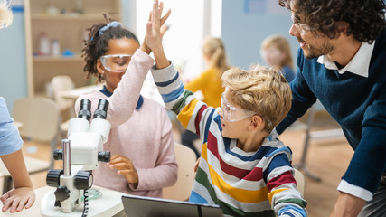 Wall Mural - Elementary School Science Classroom: Cute Little Girl Looks Uses Microscope, Boy Uses Digital Tablet Computer to Check Information on the Internet and They do High Five in Celebration.