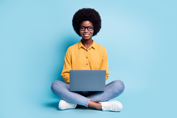 Full body size photo of black skin big volume hairstyle woman sit floor hold netbook shiny smiling cozy workplace coding website wear specs jeans yellow shirt isolated blue color background