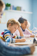 © Gorodenkoff - In Elementary School Classroom Brilliant Caucasian Boy Writes in Exercise Notebook, Taking Test and Writing Exam. Junior Classroom with Group of Children Working Diligently and Learning New Stuff
