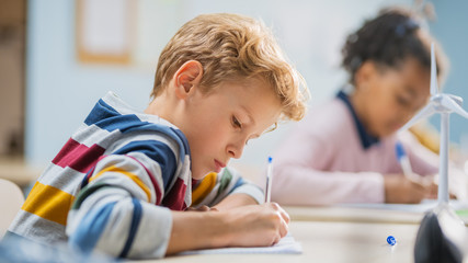 in elementary school classroom brilliant caucasian boy writes in exercise notebook, taking test and 