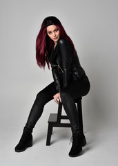 Wall Mural - Full length portrait of  girl with red hair wearing black leather jacket, pants and boots. Sitting pose on a chair, isolated against a grey studio background.