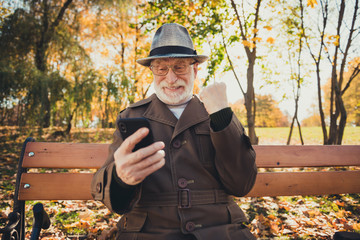 Canvas Print - Yeah october sunny weather. White grey hair old man rest relax fall town park sit bench use smartphone check internet forecast raise fists wear cap headwear hat coat jacket