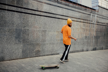  Handsome young stylish hipster guy with beard in blank orange t-shirt riding on longboard.