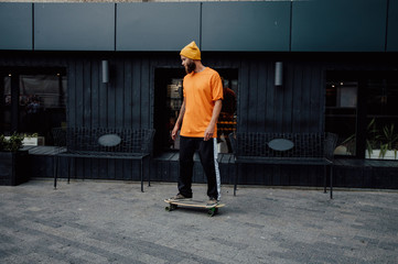 Poster - Handsome young stylish hipster guy with beard in blank orange t-shirt riding on longboard.