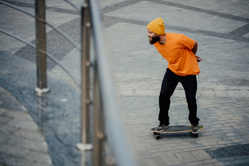 Poster - Handsome young stylish hipster guy with beard in blank orange t-shirt riding on longboard.