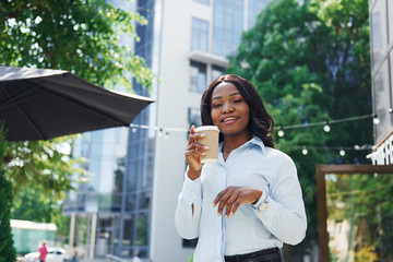 Holding cup of drink in hands. Young afro american woman in white shirt outdoors in the city near green trees and against business building