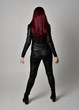 Full length portrait of  girl with red hair wearing black leather jacket, pants and boots. Standing pose facing away from the camera, isolated against a grey studio background.