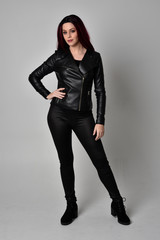 Wall Mural - Full length portrait of  girl with red hair wearing black leather jacket, pants and boots. Standing pose, isolated against a grey studio background.