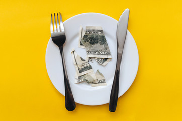 The dollar bill lies on a white plate, pierced with a fork near the knife on a yellow cotton tablecloth. The concept of business lunch, banking, rentier life and financial well-being, income reduction