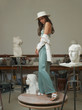 Beautiful slim woman with long dark hair in green top and palazzo trousers, white hat and oversize blazer standing at table in art studio between sculptures