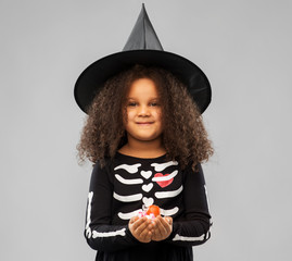 Wall Mural - halloween, holiday and childhood concept - smiling african american girl in black costume and witch hat with candies trick-or-treating over grey background