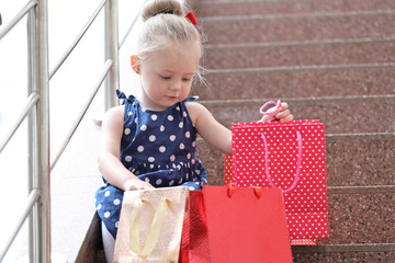 Sticker - little girl sits with colored bags on the steps in the mall.